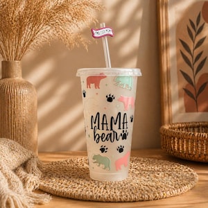 Könnte beinhalten: Transparenter Kunststoffbecher mit Deckel und Strohhalm, mit einem "Mama Bear"-Design mit Bärenillustrationen und Pfotenabdrücken. Der Strohhalm hat ein "I'M A COOL MOM"-Schild. Der Becher ist mit rosa, grünen und braunen Bären verziert.
