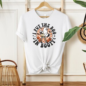 White retro ghost graphic t-shirt with orange and black checkered pattern and "I Put The Boo In Booty" text, hanging on a wooden ladder decor.
