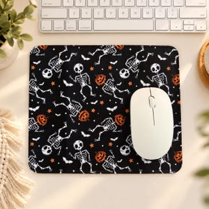 May include: A square mouse pad with a black background featuring a repeating pattern of white skeletons, orange jack-o'-lanterns, small orange stars, and black bats. A white computer mouse is positioned on the right side of the mouse pad.