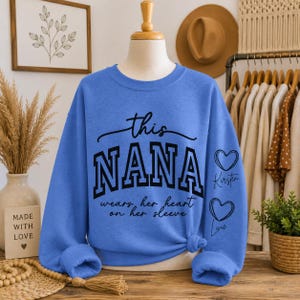 Könnte beinhalten: Ein hellblaues Sweatshirt mit dem Text "this NANA wears her heart on her sleeve" in Schwarz. Zwei Herzen mit Namen befinden sich auf dem rechten Ärmel. Das Sweatshirt wird auf einer Schaufensterpuppe präsentiert.