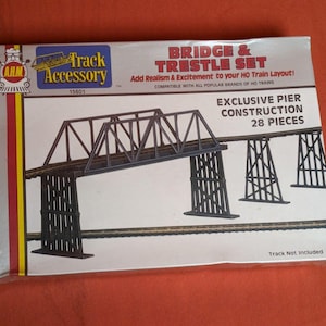 Puede incluir: Una caja para un set de puente y caballete de tren modelo a escala HO. La caja es blanca con bordes rojos y amarillos. La caja presenta una imagen del set de puente y caballete, junto con el texto "Bridge & Trestle Set", "Add Realism & Excitement to your HO Train Layout!" y "Exclusive Pier Construction 28 Pieces".