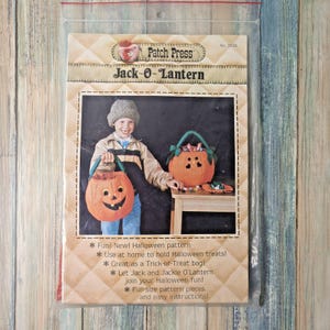 Może przedstawiać: Wzór rzemieślniczy w stylu vintage zatytułowany "Jack-O-Lantern" firmy Patch Press. Wzór przedstawia uśmiechniętą pomarańczową torbę z dyni z zielonym uchwytem, trzymaną przez dziecko. Wzór obiecuje zabawę, proste instrukcje i elementy w pełnym rozmiarze.