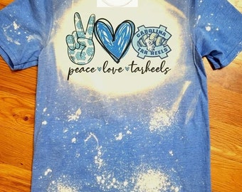 May include: A blue t-shirt with a white bleach splattered design. The shirt features a peace sign, a heart, and a Carolina Tar Heels logo with the text "peace love tarheels".