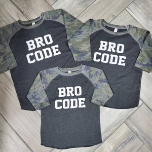 Puede incluir: Tres camisetas de béisbol negras y de camuflaje con el texto "Bro Code" en letras blancas.