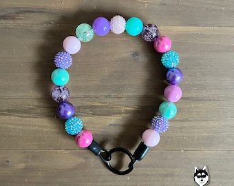 May include: A colorful beaded necklace with a black metal clasp. The beads are in shades of pink, purple, teal, and turquoise. The necklace is made of large, round beads with a sparkly finish.