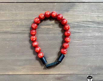 以下が含まれることがあります： 黒い留め金が付いた赤いビーズブレスレット。ブレスレットは、丸く質感のある赤いビーズでできています。留め金は黒色で、ブレスレットの両端を接続します。背景は木製の表面です。