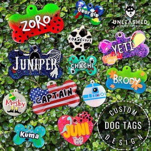 May include: A collection of custom dog tags in various shapes and colors, including bone, heart, and butterfly designs. Each tag features a unique name or design, such as "Zoro," "Juniper," and "Captain." The tags are set against a backdrop of green foliage.