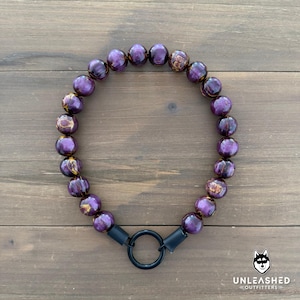 May include: A beaded dog collar featuring large, round, purple beads with gold marbling. The collar has a black metal ring and connectors. The collar is displayed on a wooden surface. The Unleashed Outfitters logo is in the bottom right corner.