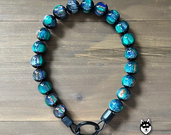 May include: A beaded dog collar featuring large, round beads in shades of teal, blue, and bronze. The beads are strung together with a black metal clasp and ring. The collar is displayed on a wooden surface. The Unleashed Outfitters logo is in the bottom right corner.