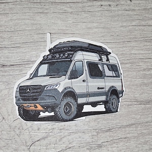 May include: A sticker featuring a white and gray van with a black roof rack, off-road tires, and a front bumper. The van has a light bar and is designed for adventure. The sticker is on a gray wood-grain background.