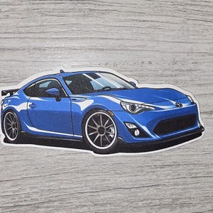 May include: A blue sports car sticker with a white border. The car has black wheels and a spoiler. The sticker is on a gray wooden surface.