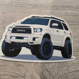 May include: White Toyota Sequoia SUV sticker with black wheels and tires. The sticker features the word "TOYOTA" on the front grill. The vehicle has a lifted suspension and a black roof rack.