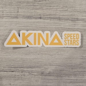 May include: A yellow sticker with the text "AKINA SPEED STARS" in white letters. The text is in a triangle shape.