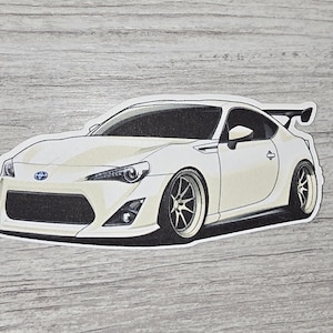 May include: A white sports car sticker with a black outline. The car has a spoiler and custom wheels. The sticker is on a gray wood-grain background.