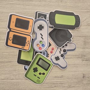 May include: A collection of colorful stickers featuring various retro video game consoles and controllers. The stickers include a green Game Boy, a black and green handheld console, a gray and white controller, and a yellow and orange handheld console.