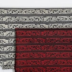 Op de afbeelding: Twee stofstalen met een herhalend patroon van zwarte en witte bloemenmotieven. De bovenste staal is zwart met een zilveren bloemenmotief. De onderste staal is rood met een zilveren bloemenmotief.