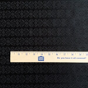 May include: Black fabric with a repeating embossed pattern of stylized flowers. A wooden ruler with the text "Do you have it all covered?" is placed on the fabric.