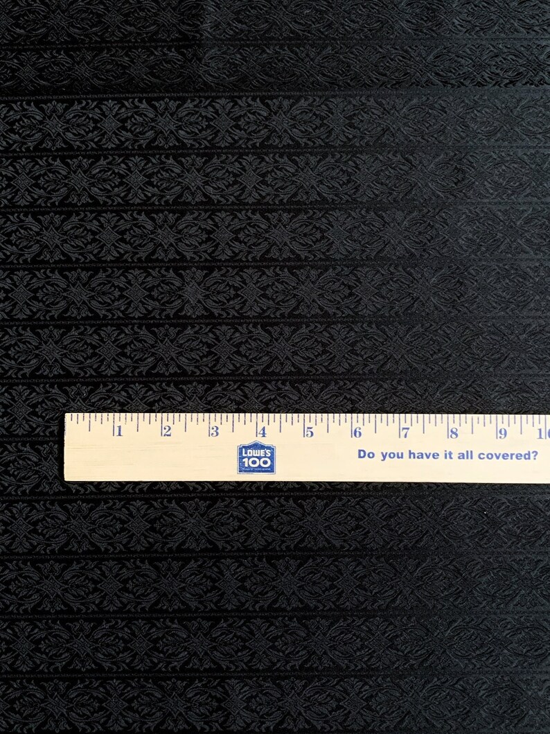 May include: Black fabric with a repeating embossed pattern of stylized flowers. A wooden ruler with the text "Do you have it all covered?" is placed on the fabric.