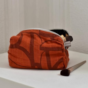 May include: An orange cosmetic bag with a dark brown abstract pattern. The bag is open, revealing makeup brushes inside. A makeup brush with a brown brush head rests on the white surface next to the bag.
