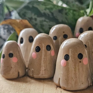 Könnte beinhalten: Eine Sammlung handgefertigter Keramik-Geisterfiguren. Jeder Geist ist hellbraun mit schwarzen Augen und rosa Wangen. Die Geister sind auf einer Holzoberfläche angeordnet, mit grünem Laub im Hintergrund.
