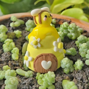 May include: A charming yellow bee figurine with a white hat and a smiling face. The bee sits on a yellow beehive decorated with white flowers and a brown door with a white heart. The figurine is surrounded by small green succulents.