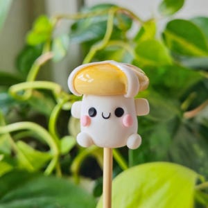 May include: A white and yellow clay mushroom with a cute face and pink cheeks, on a wooden stick.
