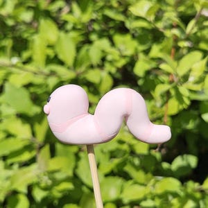 Puede incluir: Una figura en forma de gusano rosa claro con un pequeño ojo negro y detalles rosas, montada en un palito de madera. El gusano se encuentra sobre un fondo borroso de follaje verde.
