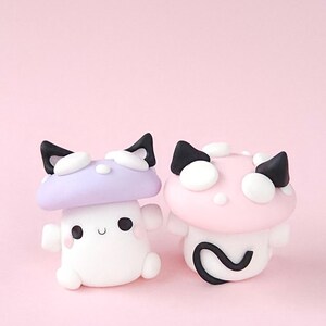 May include: Two whimsical mushroom figurines. One is white with a lavender cap and black cat ears, the other is white with a pink cap and black cat ears. Both have smiling faces and rounded bodies, set against a pink background.