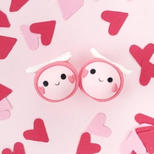 May include: Two pink clay ornaments shaped like a heart with white wings and black eyes. Each heart has a small pink heart on each side. The ornaments are on a pink background with pink hearts.