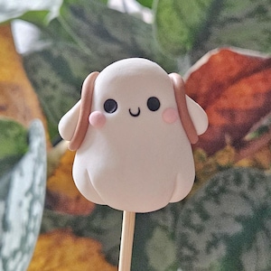 May include: A white, ghost-shaped confection on a wooden stick. The ghost has black eyes, a curved smile, and pink cheeks. It features brown ear-like accents. The background is blurred with green and orange foliage.