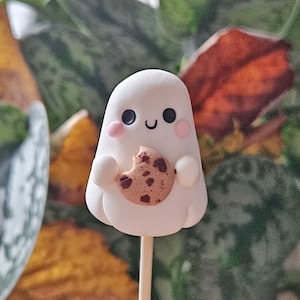May include: A white ghost-shaped cake pop with black eyes, a curved mouth, and pink cheeks, holding a chocolate chip cookie. The cake pop is on a wooden stick, set against a blurred background of green and orange foliage.