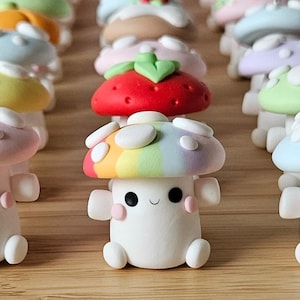 May include: A selection of charming mushroom figurines. The front-most mushroom has a rainbow-coloured cap and a smiling face. Other mushrooms have strawberry, pastel, and solid-coloured caps.