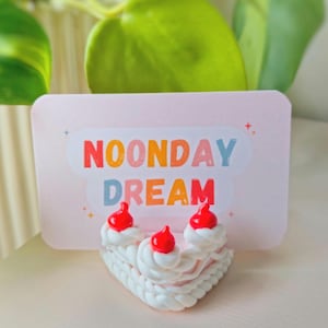 Puede incluir: Una tarjeta rosa con el texto "NOONDAY DREAM" en colores arcoíris está apoyada en un soporte blanco con forma de pastel con glaseado rojo y blanco.