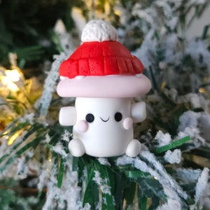 May include: A white mushroom-shaped ornament with a red and white knitted hat and a white pom-pom on top. The ornament has a smiling face with pink cheeks and is sitting on a snowy green tree branch.