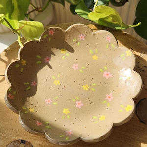 Puede incluir: Un plato de cerámica beige con borde festoneado, adornado con pequeños diseños florales rosas y amarillos. El plato está colocado sobre una superficie de madera, con follaje verde en el fondo. El plato tiene unos 20 cm de diámetro.