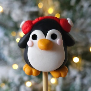 Puede incluir: Una pequeña figura de pingüino con un gorro rojo y pies naranjas, en un palo de madera. El pingüino está hecho de arcilla blanca, negra y naranja y tiene una apariencia linda y de dibujos animados.