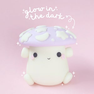Puede incluir: Un pequeño hongo blanco con un sombrero morado y manchas blancas. El hongo tiene una cara linda de dibujos animados y está etiquetado como "glow in the dark".