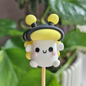 May include: A whimsical, handmade clay figurine of a smiling, cartoon-like character. The figure is primarily white with yellow accents, including two round antennae and side details. It has black eyes, a curved mouth, and pink cheeks, mounted on a wooden stick.