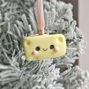 May include: A yellow cheese-shaped Christmas ornament with a smiling face, black eyes, and pink cheeks. The ornament is suspended by a light pink ribbon and is set against a snowy evergreen background.