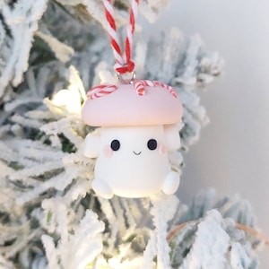 May include: A pink and white mushroom-shaped Christmas ornament with candy cane accents. The ornament has a cute cartoon face with black eyes and a smile.