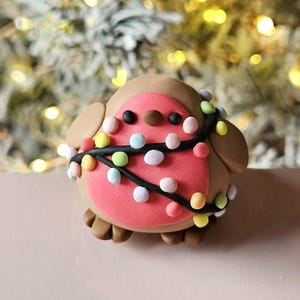 May include: A clay robin figurine with a pink breast, brown body, and a string of colourful lights wrapped around it. The bird has small black eyes and a brown beak. The background is blurred with Christmas lights.