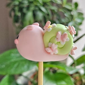 May include: A charming, pink snail-shaped cake pop with a light green shell adorned with pink butterflies and tiny white flowers. The cake pop is on a wooden stick, set against a blurred green foliage background.