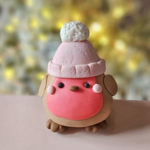 May include: A charming bird figurine, hand-sculpted with a brown body, a pink breast, and a matching pink hat topped with a white pom-pom. It has small black eyes and pink cheek details. The figurine is likely made from clay or a similar material.
