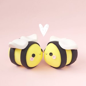 May include: Two yellow and black bumblebees with white wings are kissing. They have cute cartoon faces with pink cheeks and black eyes. A white heart is drawn above them.