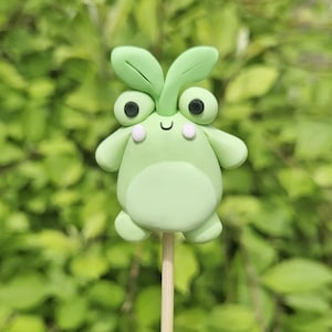 May include: A light green, handmade clay figurine of a cartoon-like creature with large eyes, small arms, and a round body. The figurine has two leaf-shaped elements on its head and is mounted on a wooden stick.