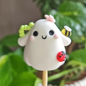 May include: A whimsical, white, ghost-shaped figurine with black eyes, a smiling mouth, and pink cheeks. Adorned with a green worm, a pink butterfly, a yellow and black bee, and a red ladybug. The figurine is mounted on a wooden stick.