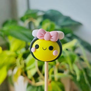 May include: A yellow and black bee cake pop with a pink bow and white antennae. The cake pop is on a wooden stick.