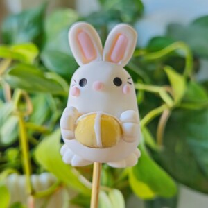 May include: A white bunny cake pop with pink ears and a yellow ball in its paws. The cake pop is on a wooden stick.