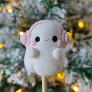 Puede incluir: Una figurita de fantasma blanco con orejeras rosas, sonriendo y con las manos delante del cuerpo. El fantasma está en un palo de madera y está delante de un fondo borroso de un árbol de Navidad con luces.