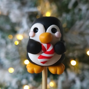 May include: A charming penguin figurine, featuring a black and white body, orange feet and beak, and holding a red and white striped candy cane. The penguin is mounted on a wooden stick, ideal for a cake topper or festive decoration.